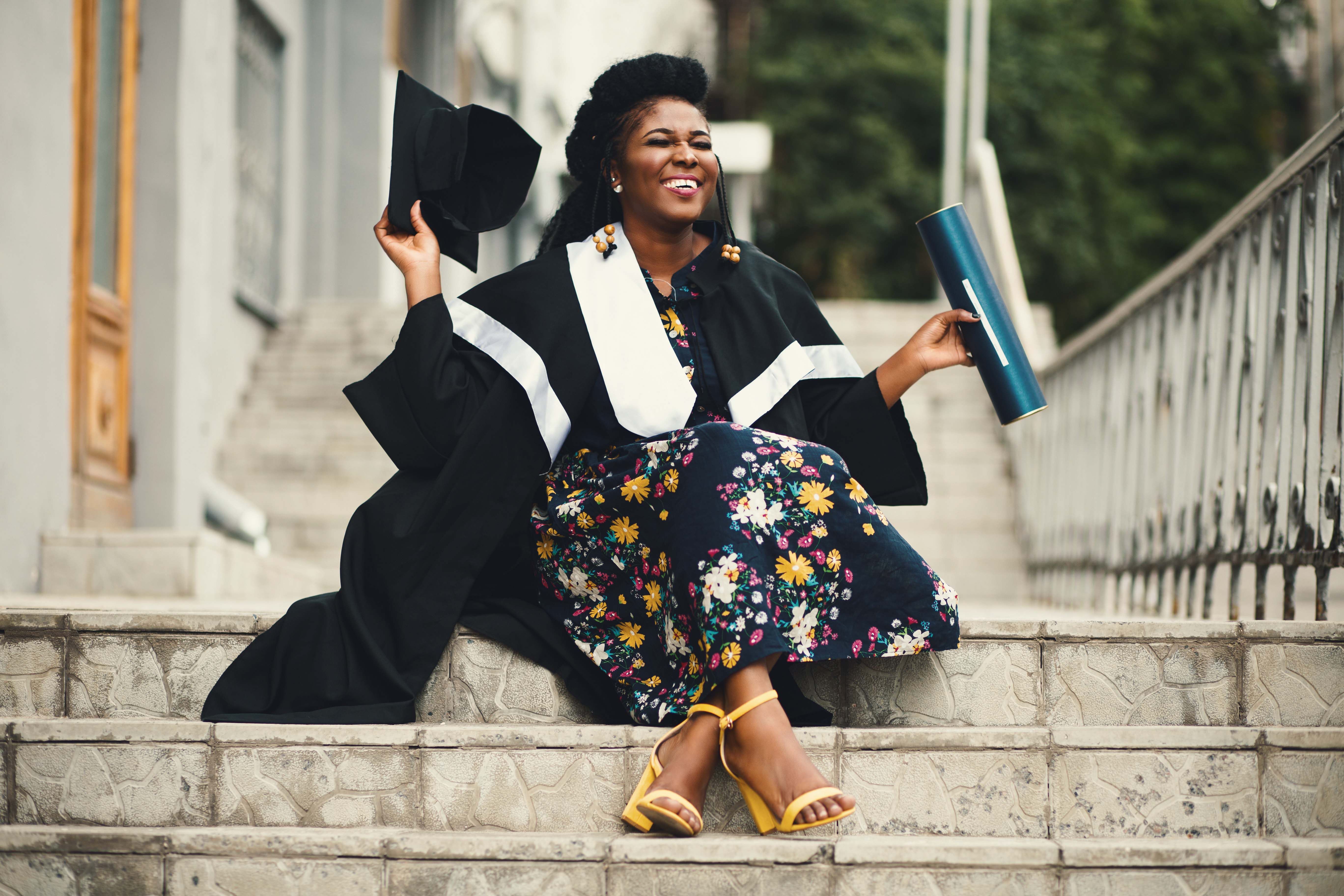 A smiling graduate sits on the steps of a campus building